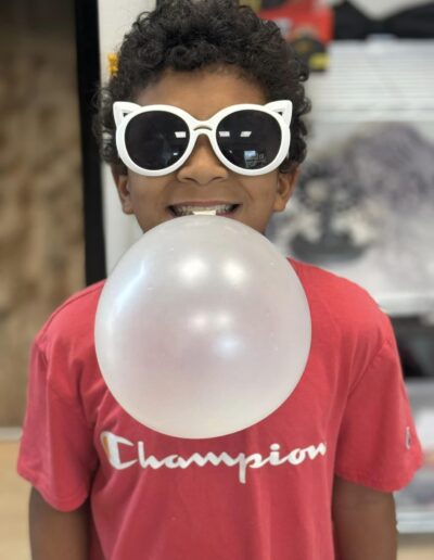 Child smiling with balloon in mouth wearing sunglasses.